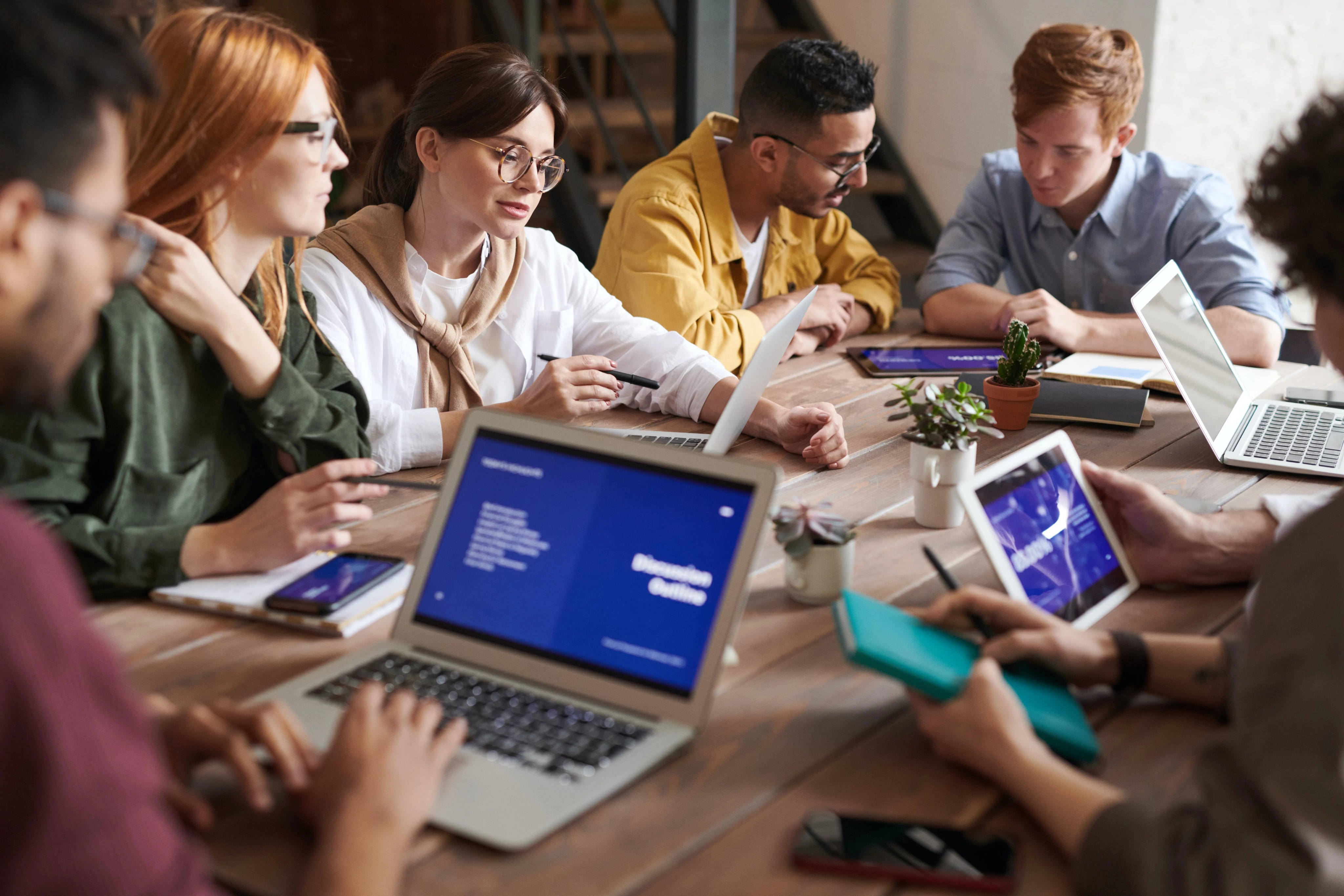 A group of six people working with laptops and tablets around a wooden table.