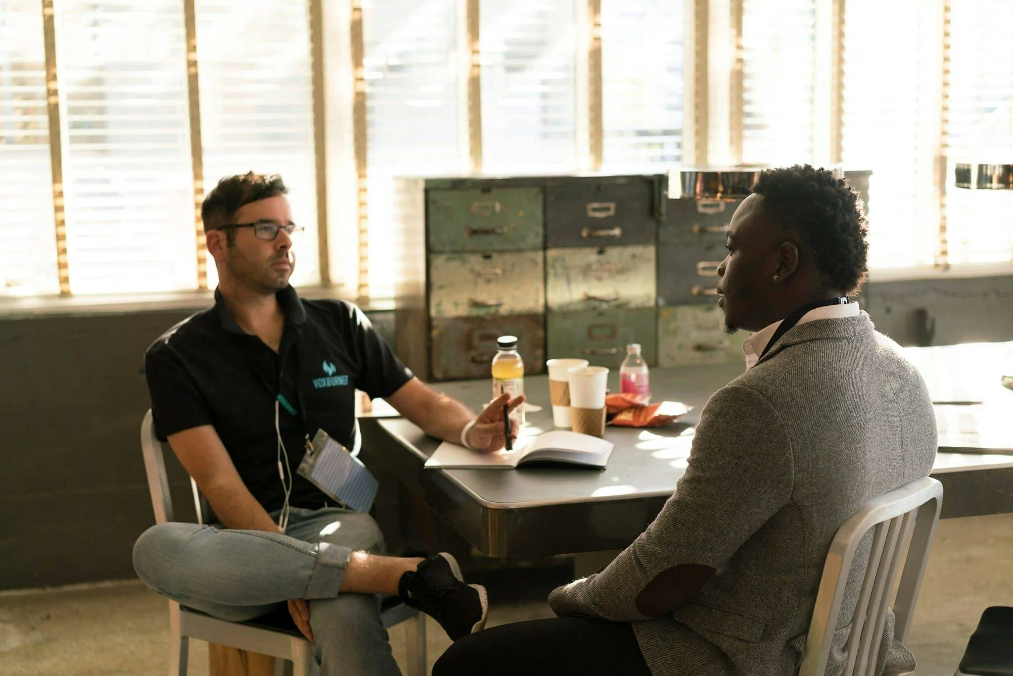 Two people sit at a table in discussion, sunlight filters through blinds.