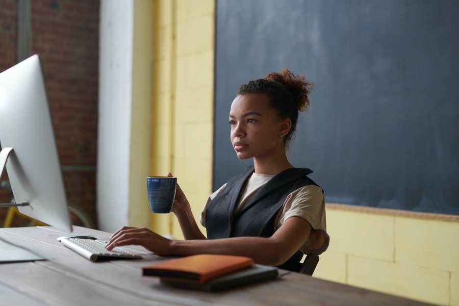 Woman at a desk with a computer, holding a mug, in a classroom setting.